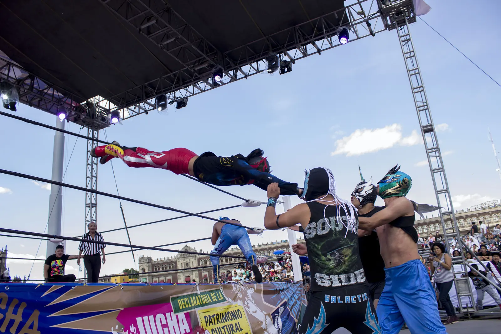 Luchador v červeném a černém skáče přes lana ringu pod širým nebem na soupeře, zatímco maskovaní zápasníci přihlížejí pod modrým nebem