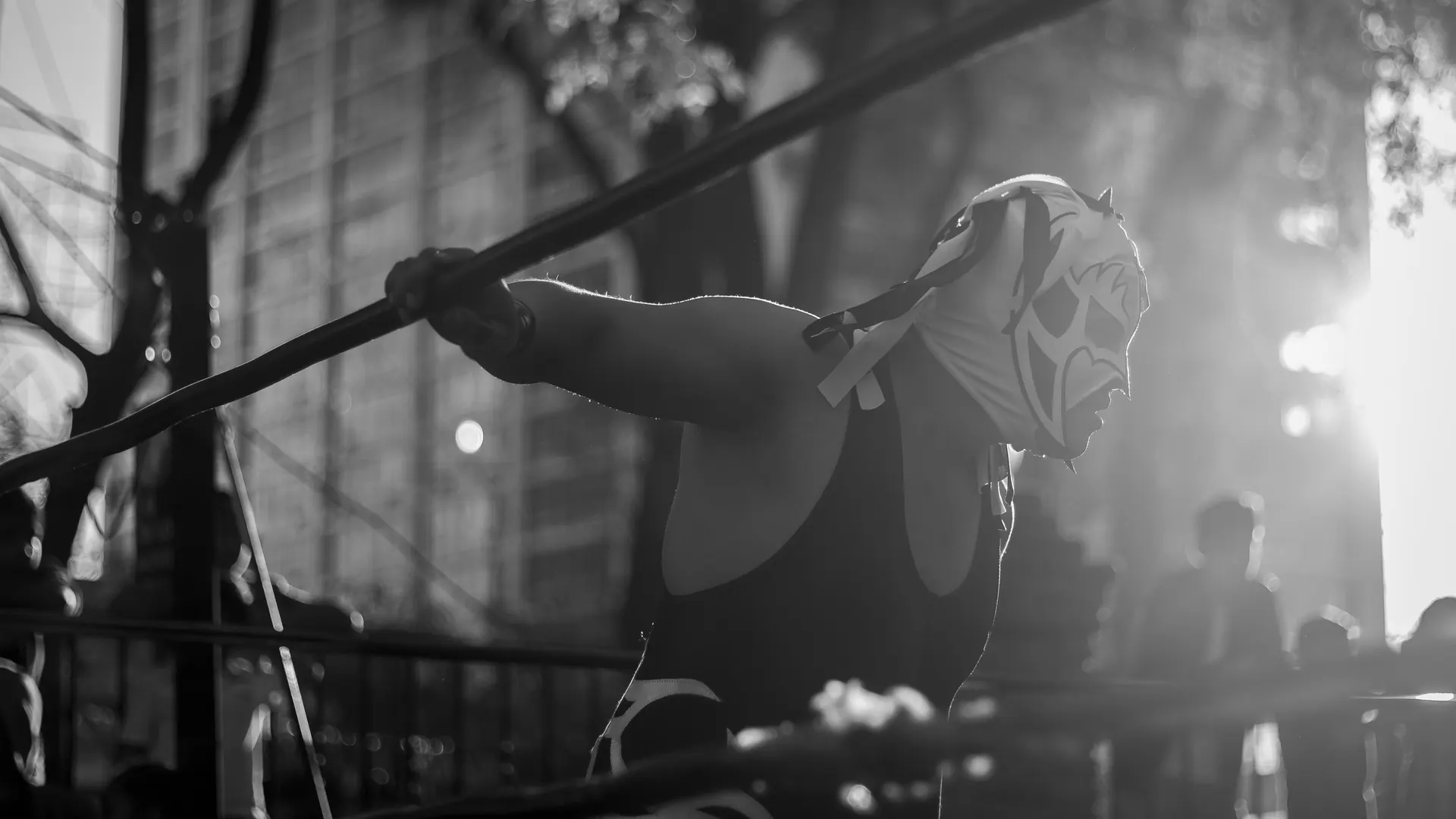 Maskovaný luchador v černém trikotu se opírá o lana ringu, podsvícený nízkým sluncem, na černobílé fotografii