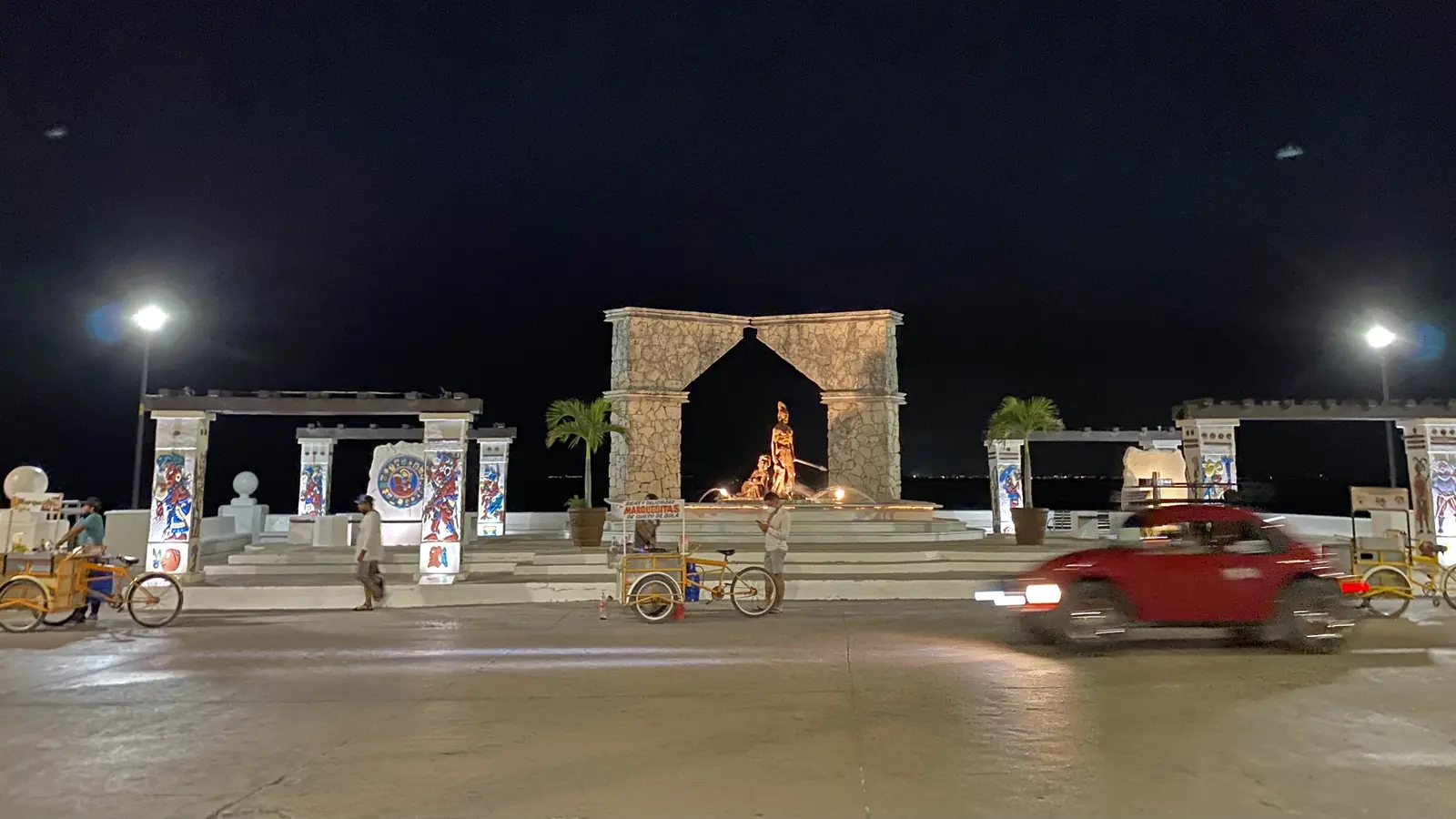 Kamenný oblouk a nasvícené památky na nábřeží San Miguel de Cozumel v noci, kolem projíždí červené auto a na náměstí stojí zaparkované tříkolky