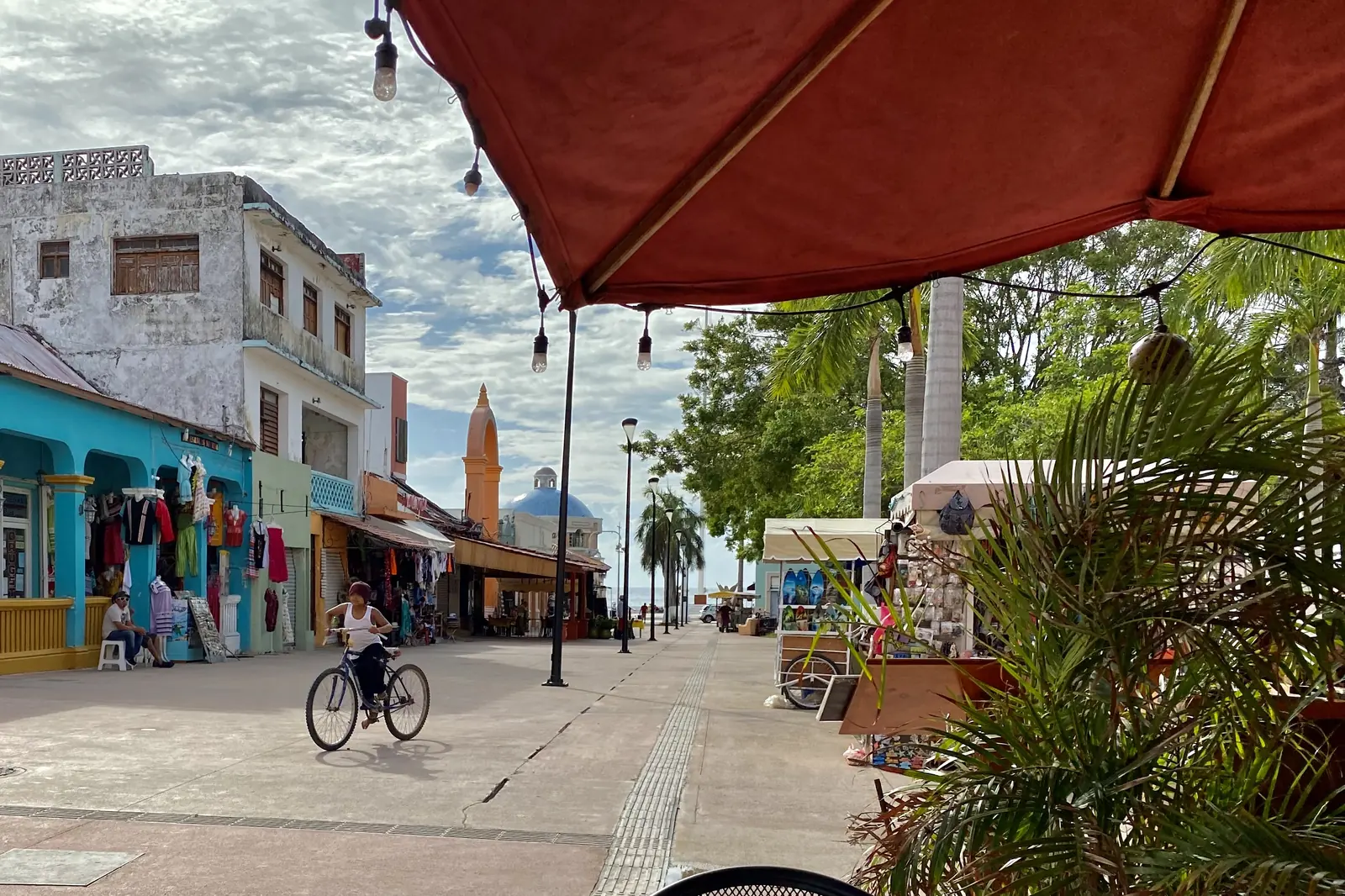 Cyclista projíždí po ulici San Miguel de Cozumel s barevnými výlohami pod markýzou, v dálce se tyčí kostelní věž