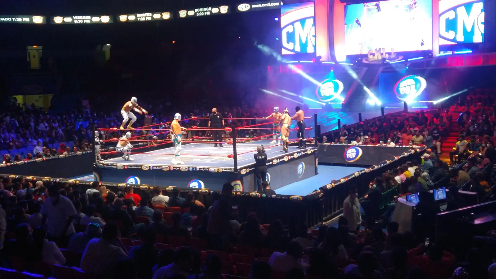 Μια φωτισμένη πίστα lucha libre σε μια μεγάλη αρένα, με luchadores στην πίστα, έναν διαιτητή και μεγάλες οθόνες βίντεο από πάνω